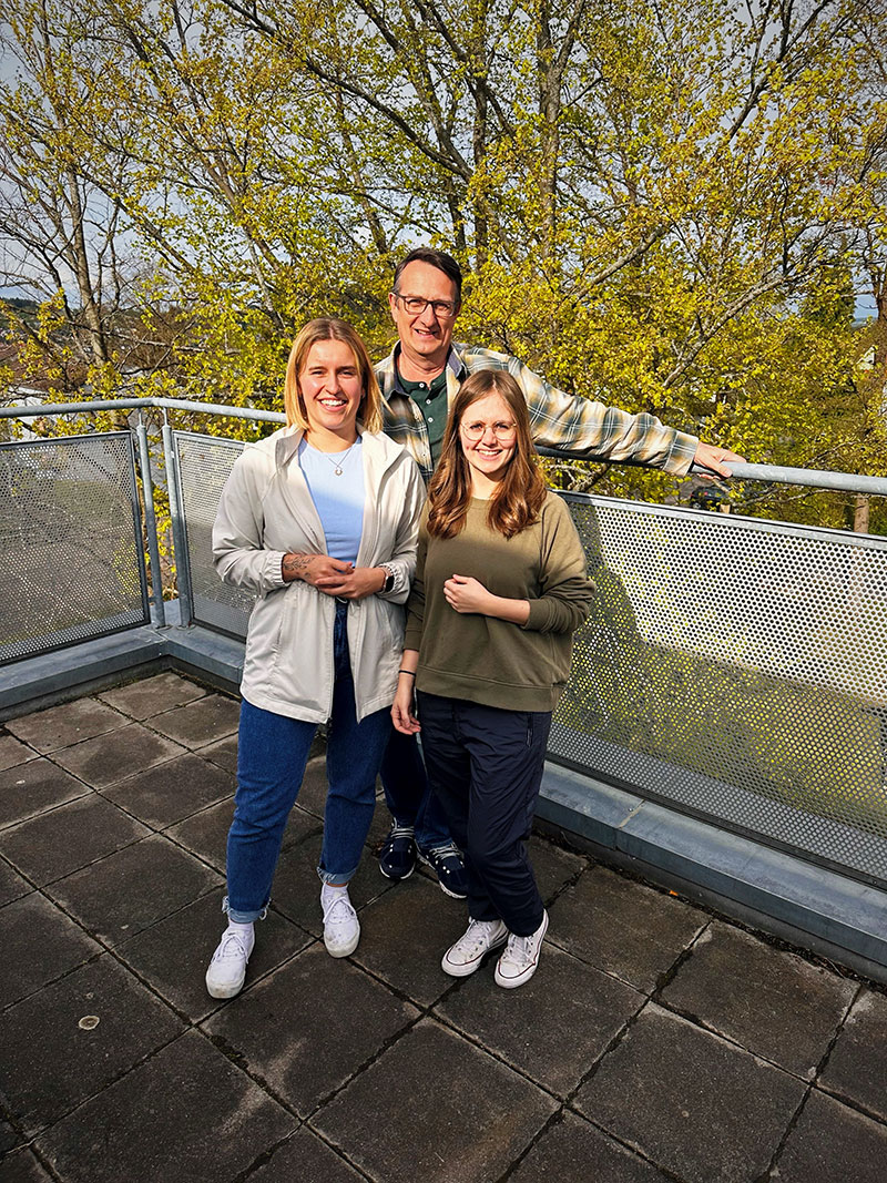 Ein Mann auf einem Balkon, vor ihm stehen zwei junge Frauen. Sie lächeln.