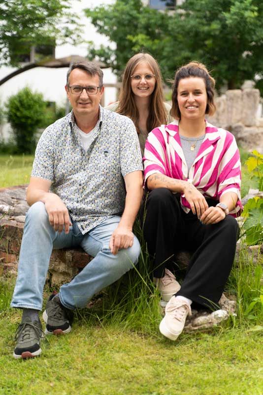 Zwei junge Frauen und ein Mann sitzen im Garten und lächeln in die Kamera
