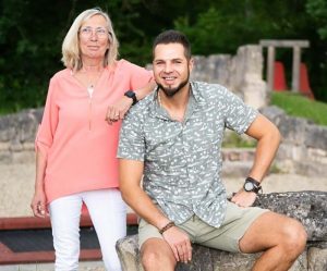 Ein Mann sitzt auf einer Steinmauer, eine Frau steht hinter ihm und legt den Arm auf seine Schulter.